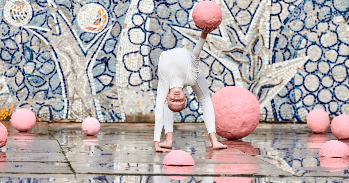Outdoor dance of young ballerina girl with alopecia in white futuristic suit with plastic and flexible movements among pink spheres on abstract mosaic Soviet background, symbolizes self expression